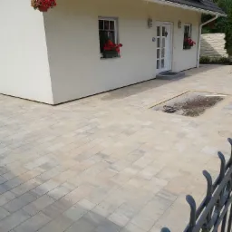 Jasna kostka brukowa ułożona przed domem z białymi oknami i czerwonymi kwiatami w donicach, widoczne puste miejsca przygotowane pod nasadzenia roślin, ogrodzenie z kutego żelaza na pierwszym planie.