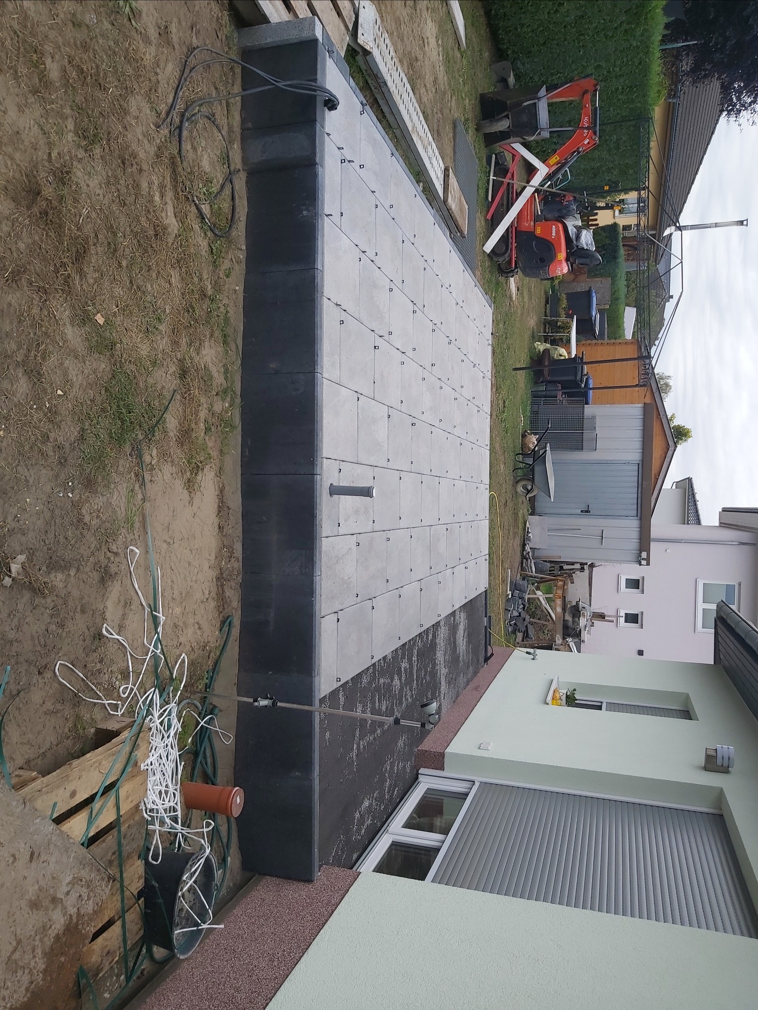 Płyty tarasowe ułożone na czarnej hydroizolacji, widoczny mini-koparka w tle, przygotowania do dalszych prac wykończeniowych.