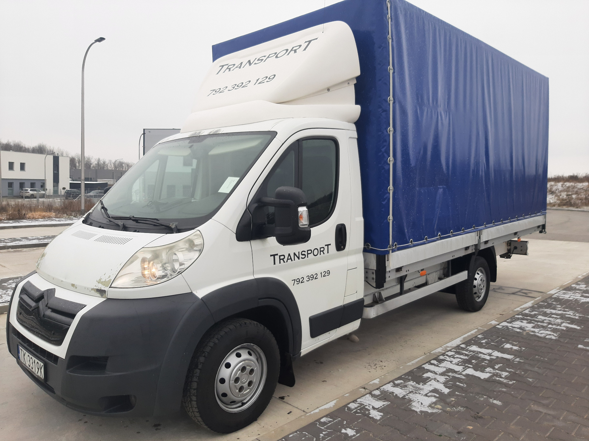 Biały bus z niebieską plandeką zaparkowany na betonowym placu, widoczne logo i numer telefonu firmy.