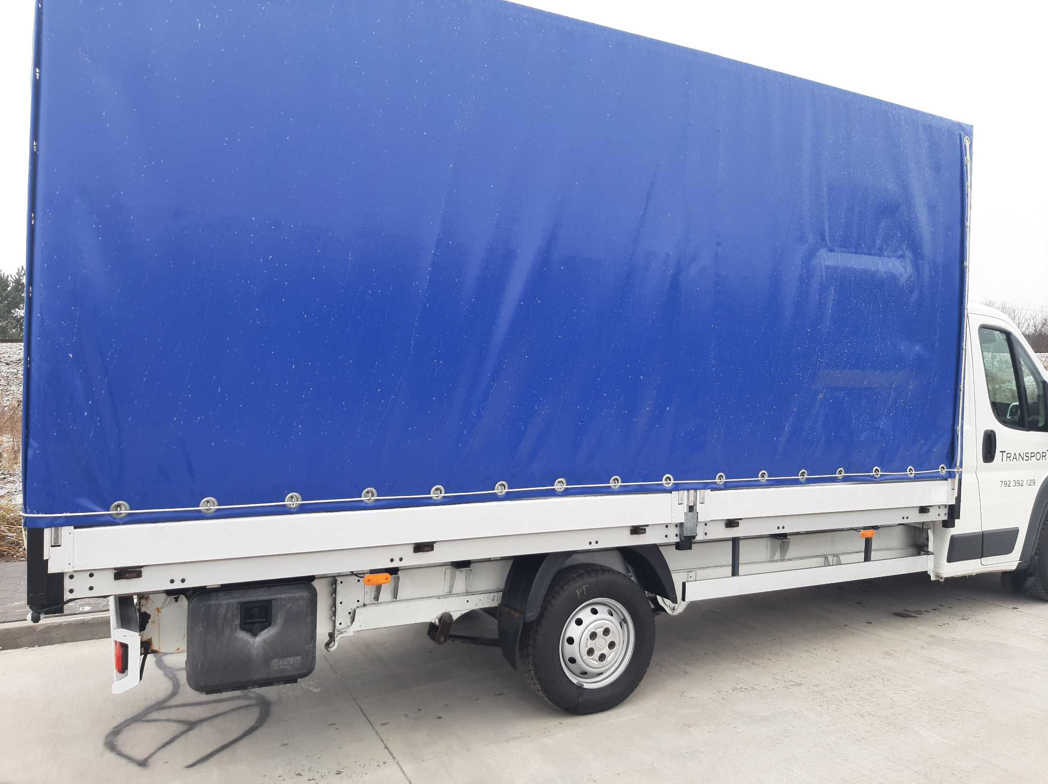 Biały bus transportowy z niebieską plandeką, widoczne mocowania plandeki oraz skrzynka narzędziowa pod spodem, na betonowym podłożu.