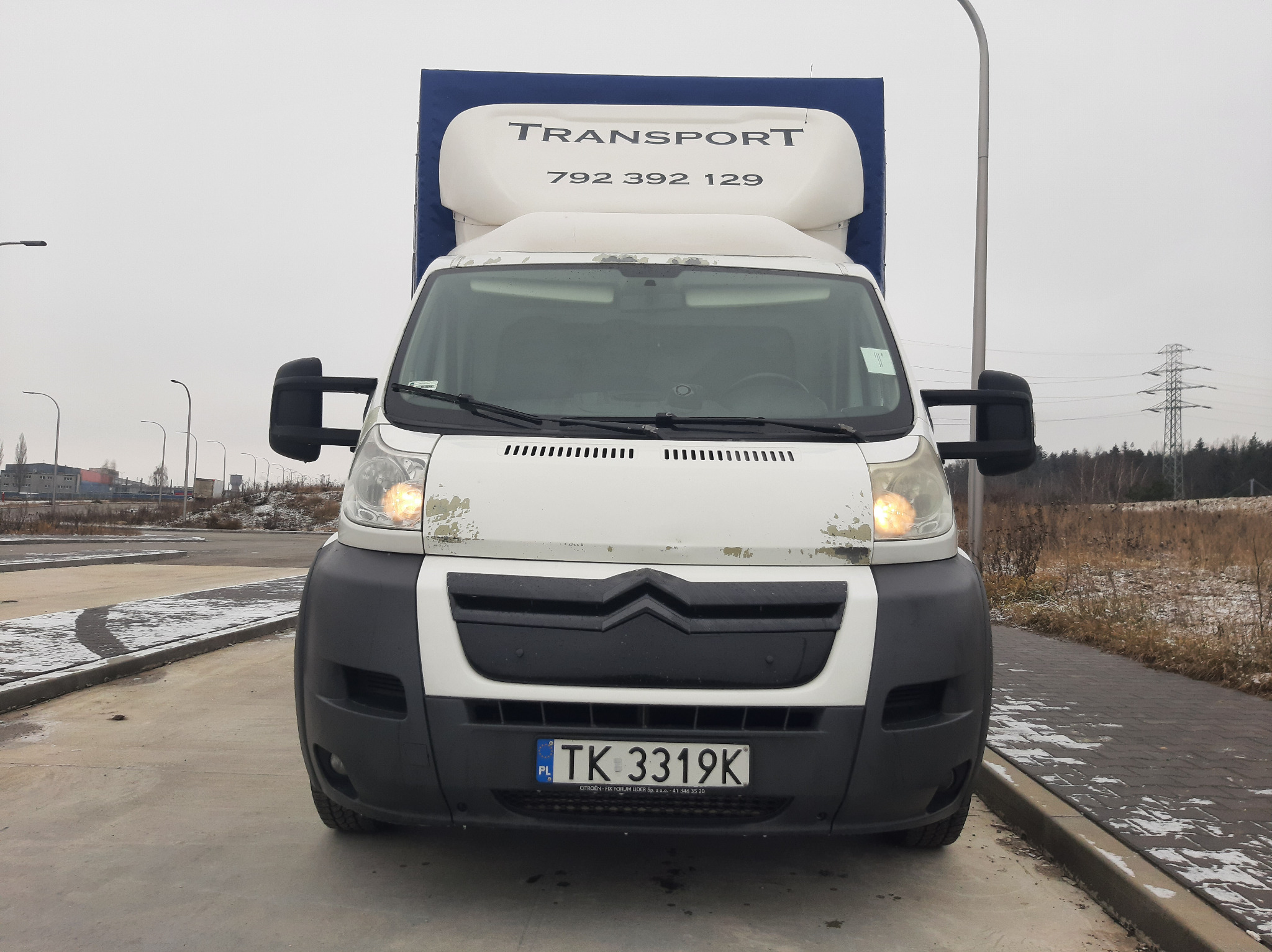 Biały samochód dostawczy z niebieską plandeką i napisem TRANSPORT oraz numerem telefonu, widziany z przodu na betonowym placu w pochmurny dzień.