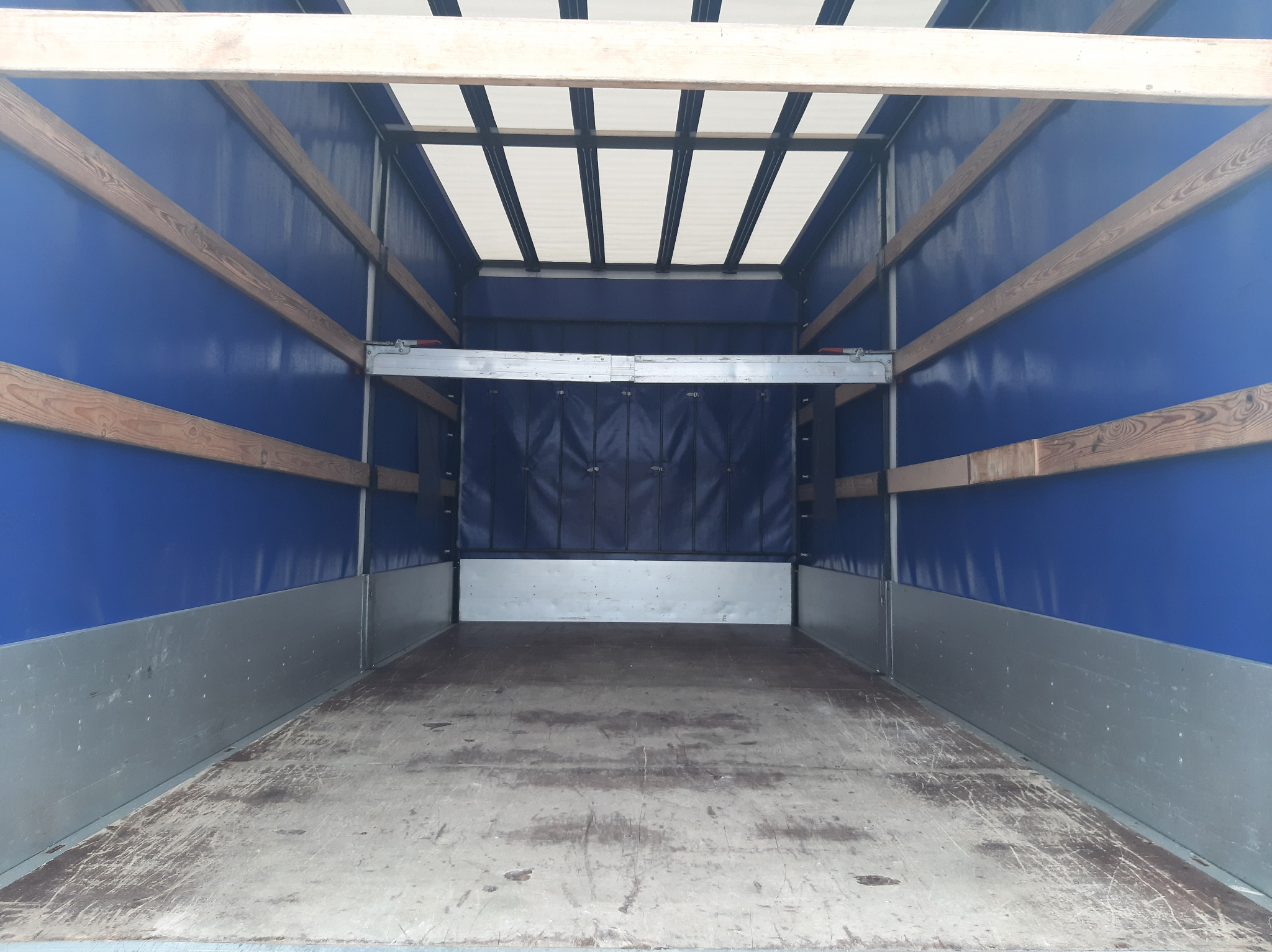 Pusta przestrzeń ładunkowa wewnątrz pojazdu dostawczego z drewnianą podłogą, niebieskimi ścianami z drewnianymi wzmocnieniami i białym dachem, gotowa do załadunku towarów.