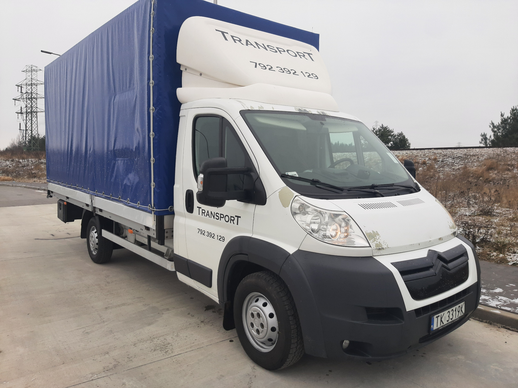 Biały samochód dostawczy z plandeką, z numerem telefonu na nadbudówce, zaparkowany w Kielcach na betonowym placu w pochmurny dzień. Widoczne drzewa w tle.