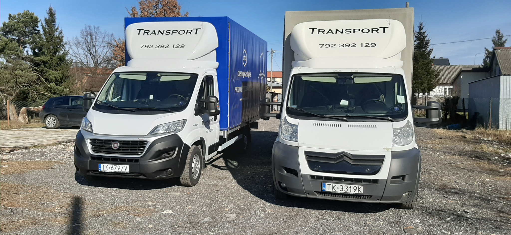 Dwa białe busy, jeden z plandeką w kolorze niebieskim z nadrukowanym napisem 'chemiplastyka' oraz numerem telefonu, zaparkowane na żwirowym placu w słoneczny dzień.