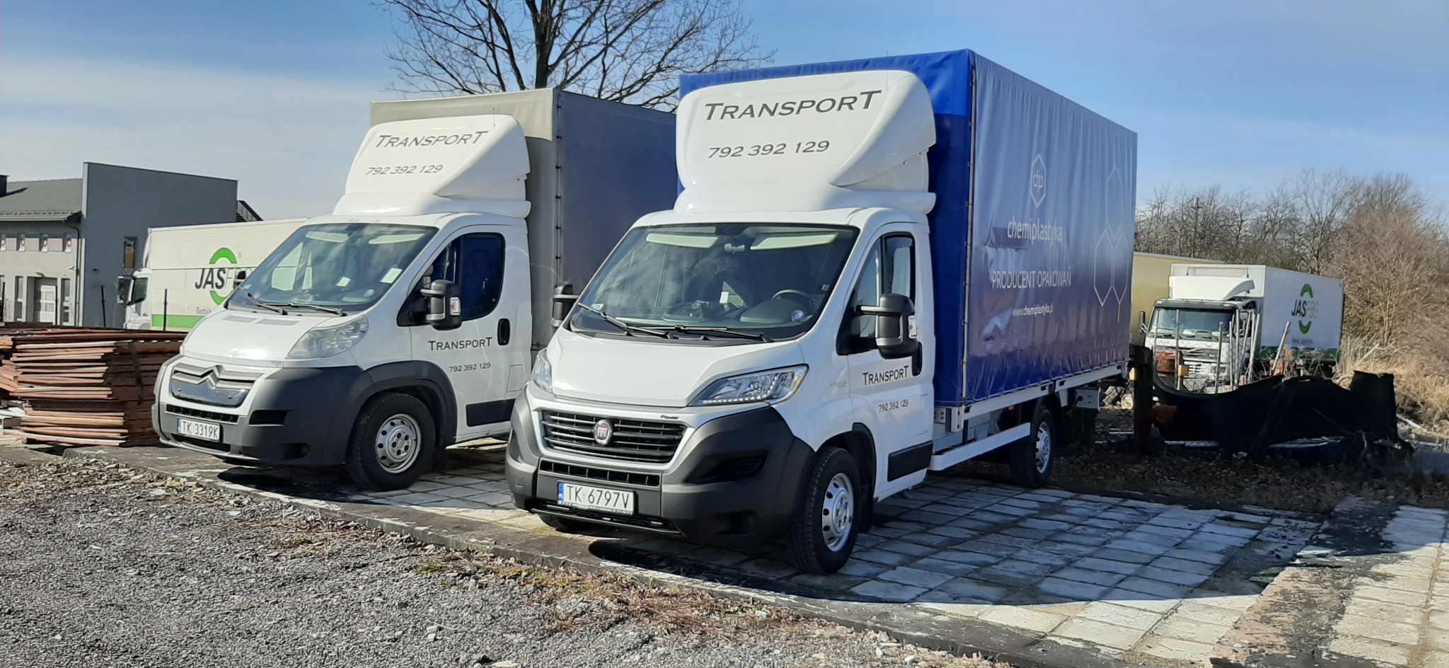 Dwa busy transportowe, biały Citroen i Fiat Ducato z niebieską plandeką, stoją zaparkowane na brukowanym placu w słoneczny dzień. Na plandece Ducato widoczne logo firmy chemiplastyka. W tle inne...