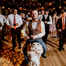 Animator w kamizelce i muszce na kostiumie 'na barana' z dużą głową, prowadzący zabawę taneczną dla gości weselnych w sali z drewnianą konstrukcją i ciepłym oświetleniem.