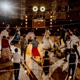 Parkiet pełen tańczących gości weselnych, oświetlony dekoracyjnymi żarówkami i reflektorami, w tle stanowisko DJ'a z podświetlonym napisem 'DJ Contigo'.