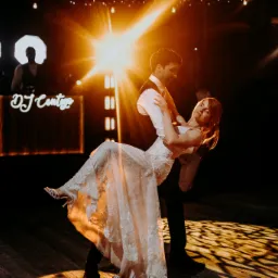 Para młoda tańcząca pierwszy taniec na parkiecie, oświetlona ciepłym światłem reflektorów, w tle widoczny DJ z podświetlonym stanowiskiem.