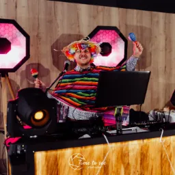 Uśmiechnięty mężczyzna w kolorowym ponczo, słomkowym kapeluszu z kwiatami i z marakasami w dłoniach, stoi za konsoletą DJ-a otoczony przez oświetlenie sceniczne.