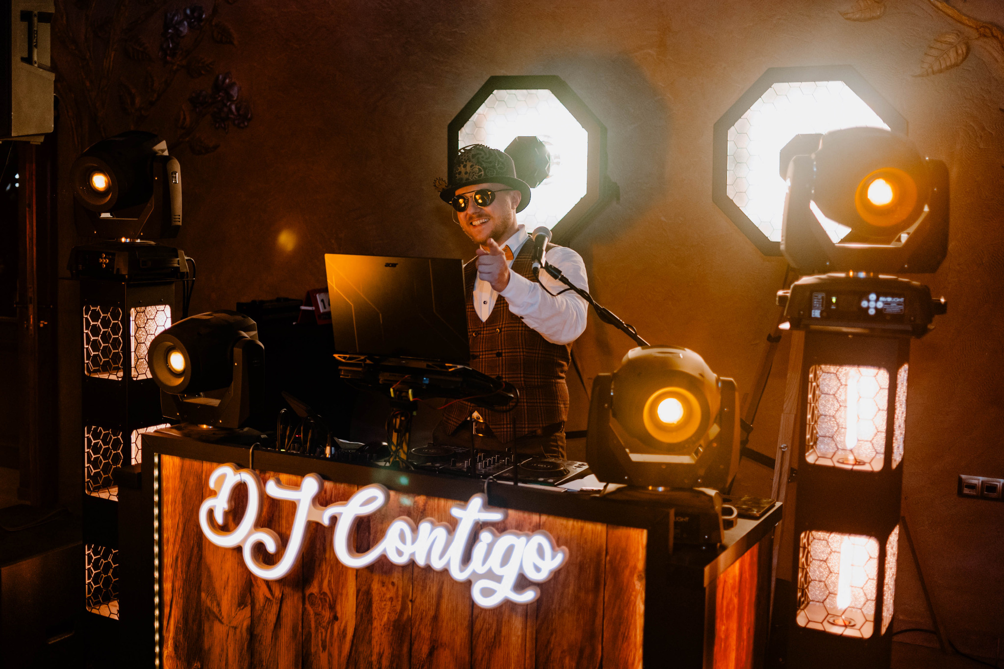 Uśmiechnięty DJ w cylindrze i okularach, wskazujący palcem, za konsoletą z napisem 'DJ Contigo', otoczony profesjonalnym oświetleniem scenicznym.