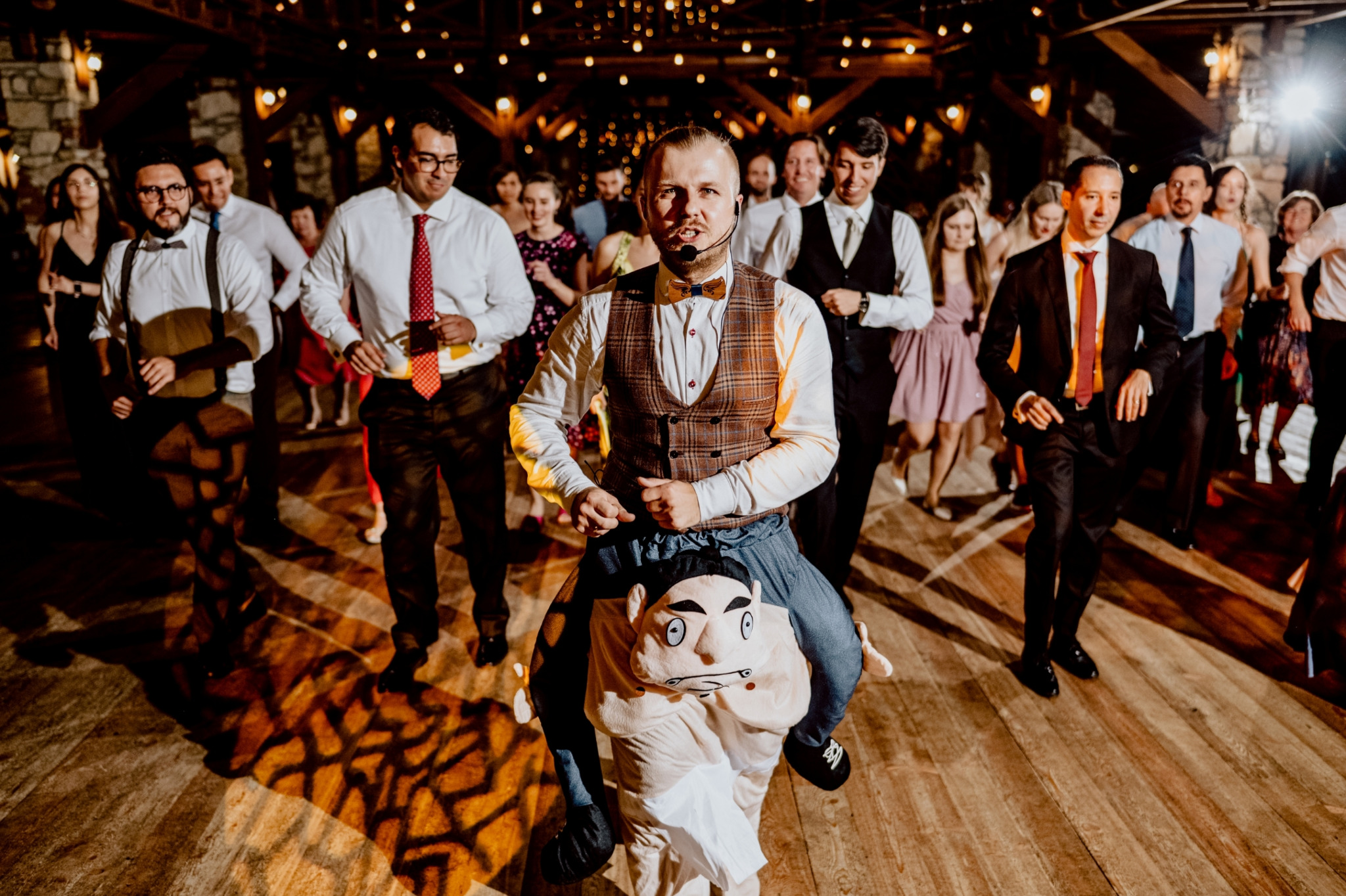 Animator w kamizelce i muszce na kostiumie 'na barana' z dużą głową, prowadzący zabawę taneczną dla gości weselnych w sali z drewnianą konstrukcją i ciepłym oświetleniem.