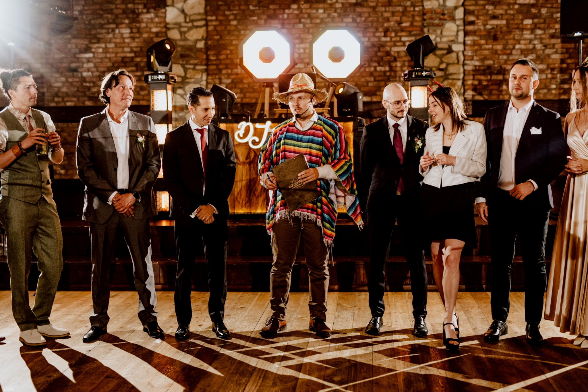 Grupa osób, w tym mężczyzna w kolorowym ponczo i kapeluszu, stoi na scenie przed podświetlonym napisem 'DJ', prawdopodobnie podczas uroczystości lub imprezy.