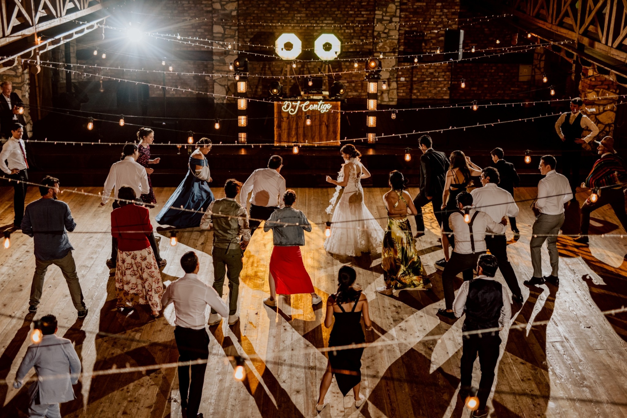 Parkiet pełen tańczących gości weselnych, oświetlony dekoracyjnymi żarówkami i reflektorami, w tle stanowisko DJ'a z podświetlonym napisem 'DJ Contigo'.