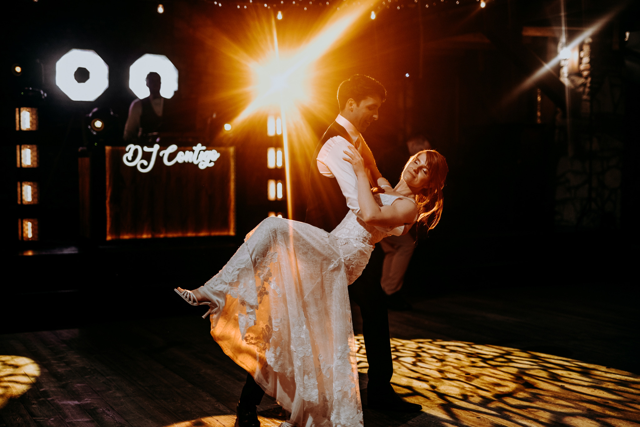 Para młoda tańcząca pierwszy taniec na parkiecie, oświetlona ciepłym światłem reflektorów, w tle widoczny DJ z podświetlonym stanowiskiem.