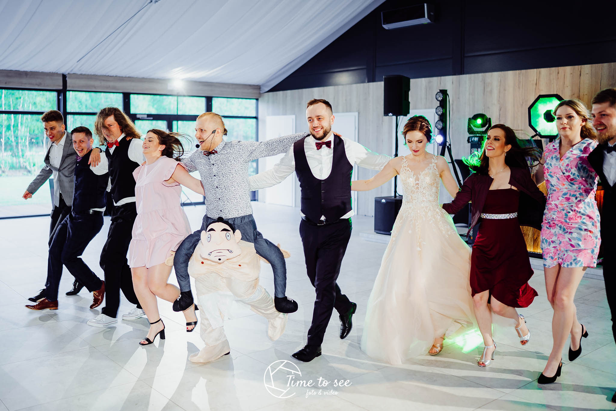 Uczestnicy wesela tańczą w korowodzie, pierwszy mężczyzna niesie na barana osobę w kostiumie karykaturalnej postaci, mikroport na głowie, para młoda w eleganckich strojach, oświetlenie sceniczne...