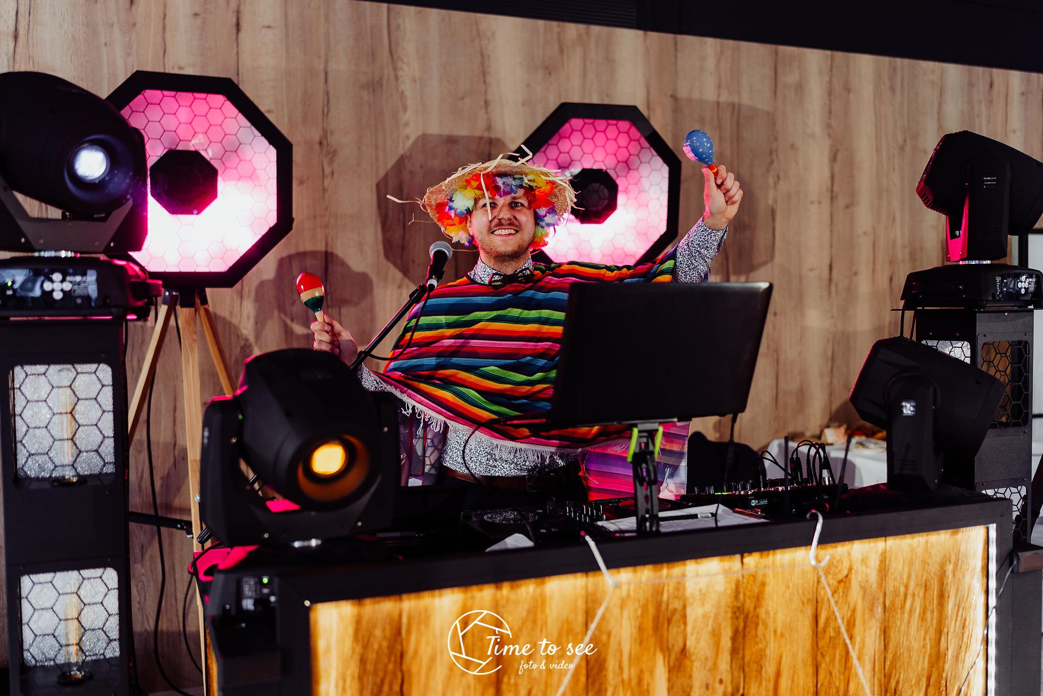 Uśmiechnięty mężczyzna w kolorowym ponczo, słomkowym kapeluszu z kwiatami i z marakasami w dłoniach, stoi za konsoletą DJ-a otoczony przez oświetlenie sceniczne.