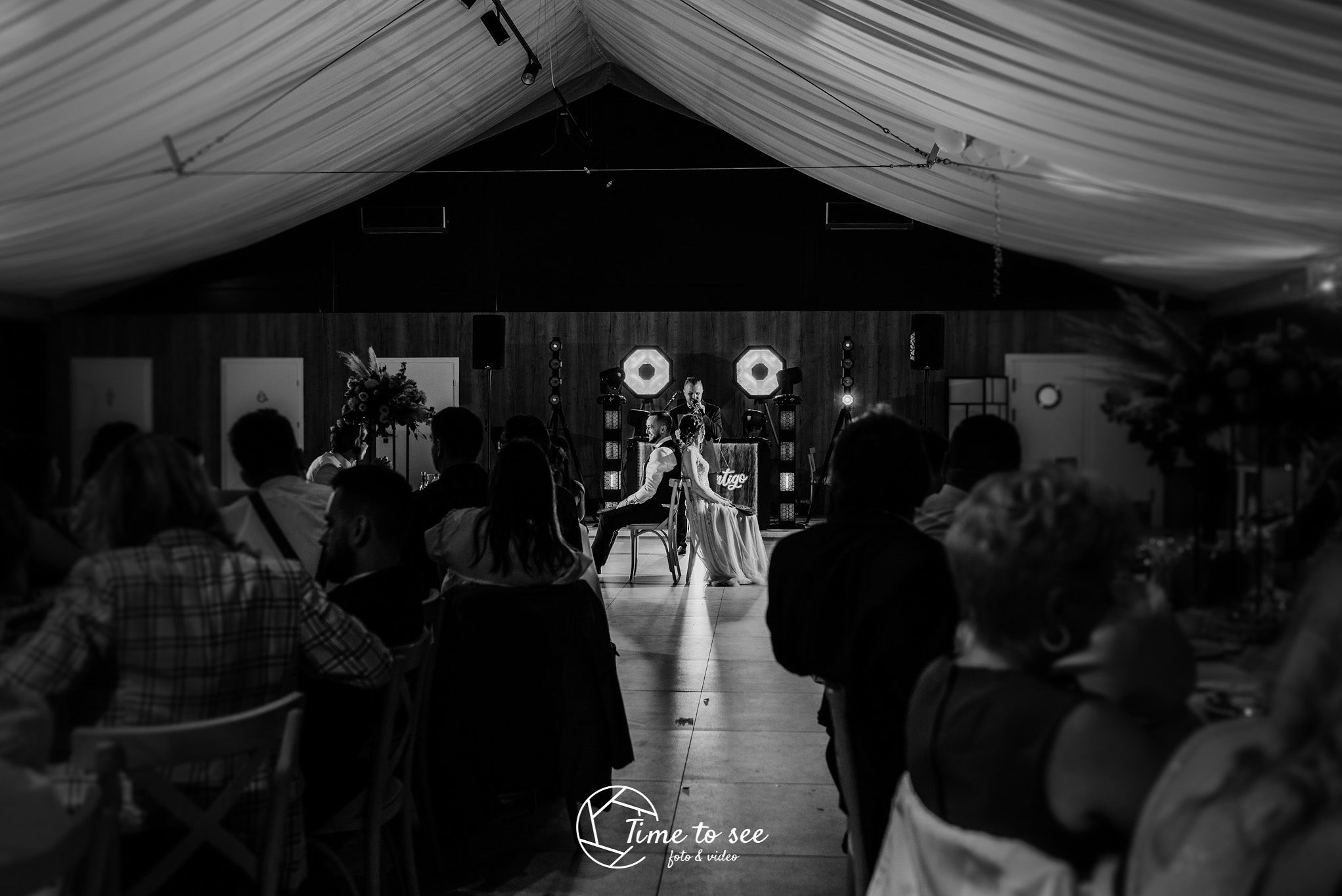 Czarno-białe zdjęcie pary młodej siedzącej na krzesłach tyłem do widowni, z DJ-em i oświetleniem scenicznym w tle, widok z perspektywy gości weselnych w sali z dekoracyjnym sufitem.