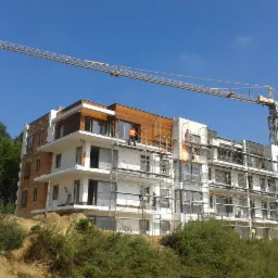 Budynek wielorodzinny w trakcie budowy z widocznym żurawiem i pracownikami na rusztowaniach, częściowo wykończony drewnianą elewacją, na tle błękitnego nieba.