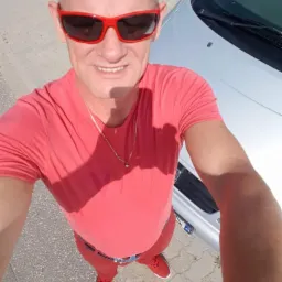 Mężczyzna w czerwonym ubraniu i okularach stoi obok srebrnego samochodu, patrząc w górę w obiektyw aparatu. Nietypowy kadr z dołu.