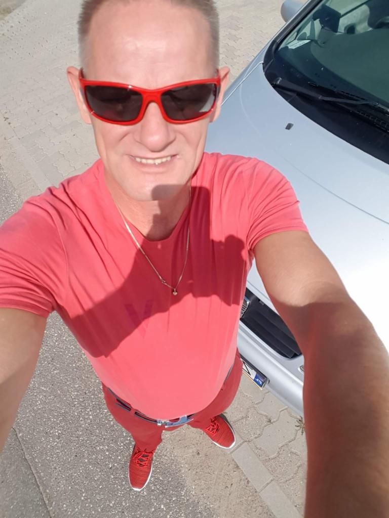 Mężczyzna w czerwonym ubraniu i okularach stoi obok srebrnego samochodu, patrząc w górę w obiektyw aparatu. Nietypowy kadr z dołu.