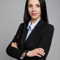 Portret kobiety w czarnym garniturze i błękitnej koszuli, z zegarkiem na lewym nadgarstku i pierścionkami, stojącej na szarym tle z założonymi rękami.