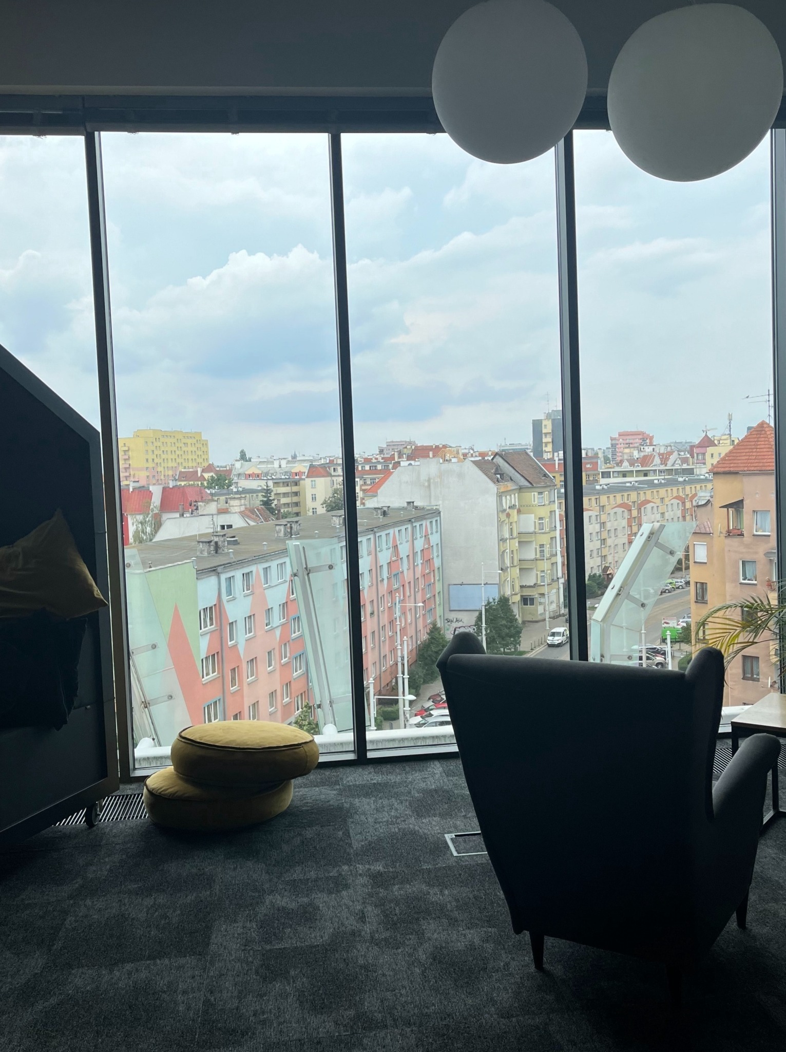 Wnętrze biura z widokiem na miasto przez duże okno; fotel z wysokim oparciem, dwa okrągłe pufy i lampy kuliste pod sufitem.