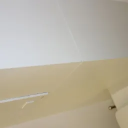 Wykończona łazienka z białymi płytkami na ścianach i podwieszanym sufitem w kolorze kremowym, widoczny przycisk spłuczki wbudowany w ścianę i fragment toalety.