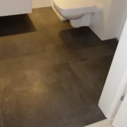 Ciemnobrązowe, kwadratowe płytki podłogowe w łazience, widok z perspektywy wejścia, z widoczną białą toaletą wiszącą i fragmentem białej szafki pod umywalką.