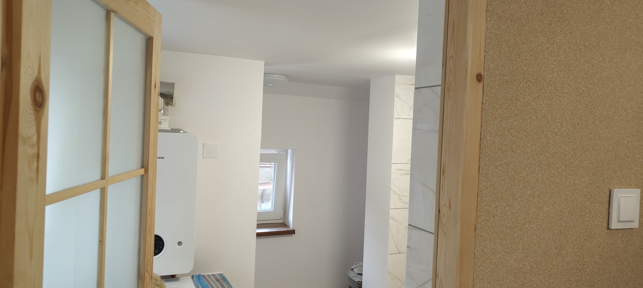 Jasne wnętrze łazienki po remoncie: białe ściany, drewniane drzwi z matowym szkłem, piec gazowy i okno. Widoczna ściana wykończona korkiem.