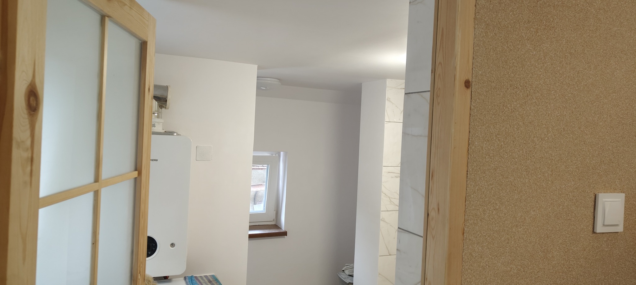 Nowoczesna łazienka po remoncie: białe ściany, imitacja marmuru na kolumnie, okno i drewniane drzwi z matowym szkłem. Na ścianie piec gazowy i włącznik światła.