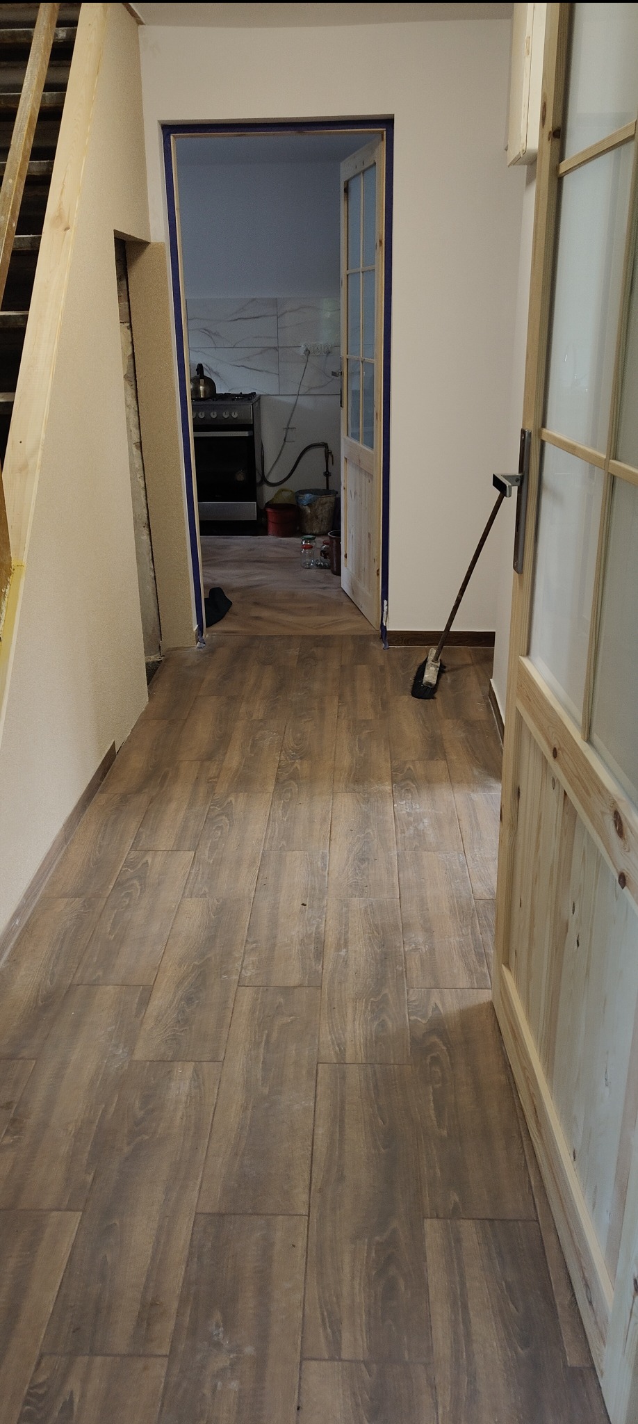 Ułożona podłoga z paneli imitujących drewno w przedpokoju, widok na otwarte drzwi do kuchni. Widoczna szczotka oparta o framugę.