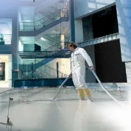 Pracownik w białym kombinezonie i żółtych kaloszach rozprowadza płynną masę na podłodze w przestronnym, nowoczesnym wnętrzu z widocznymi schodami i elementami szklanymi.