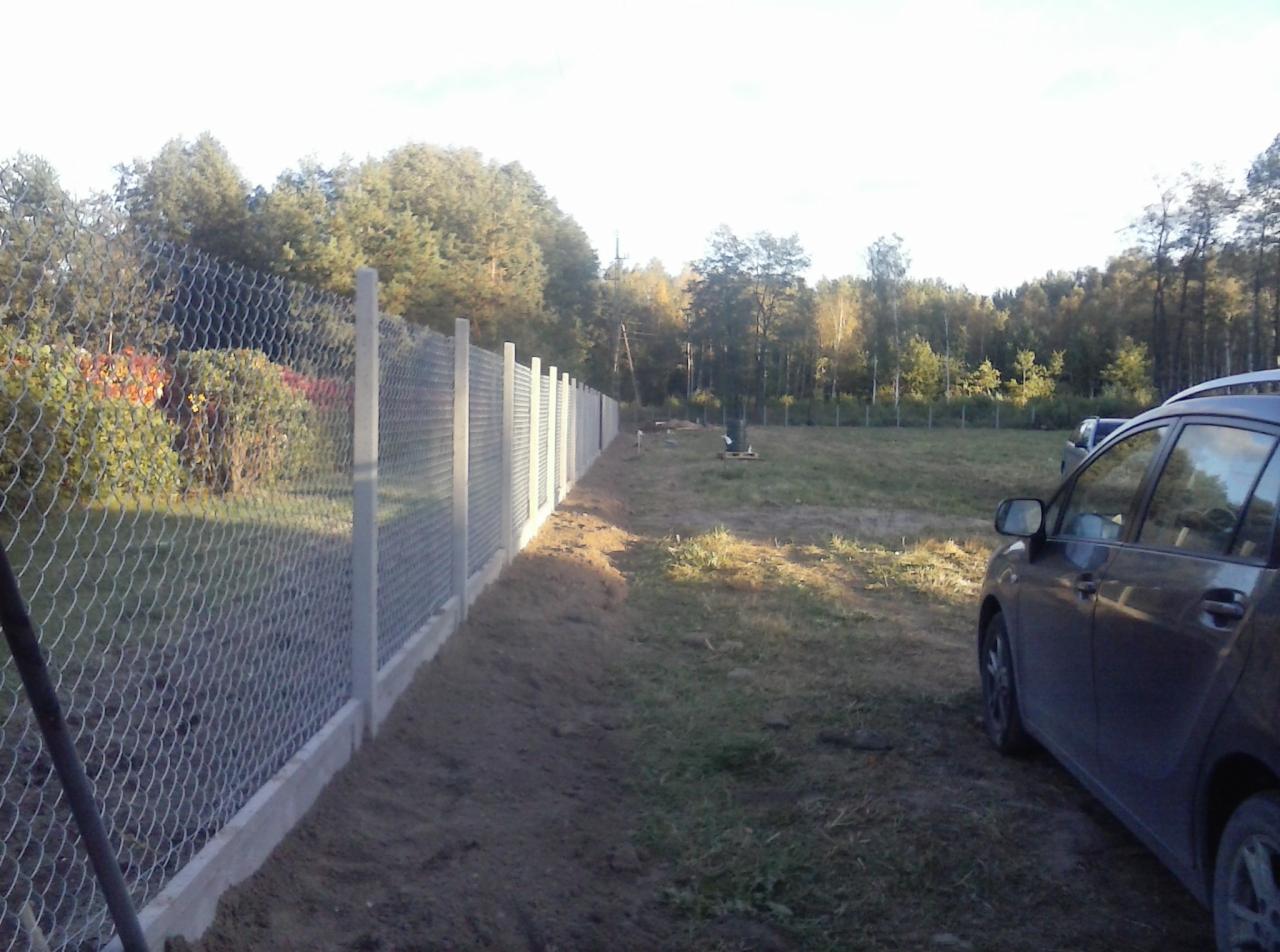 Długi, siatkowy płot z betonowymi słupkami i podmurówką, rozciągający się wzdłuż granicy działki z widocznym samochodem w tle, na tle lasu w jesiennej scenerii.