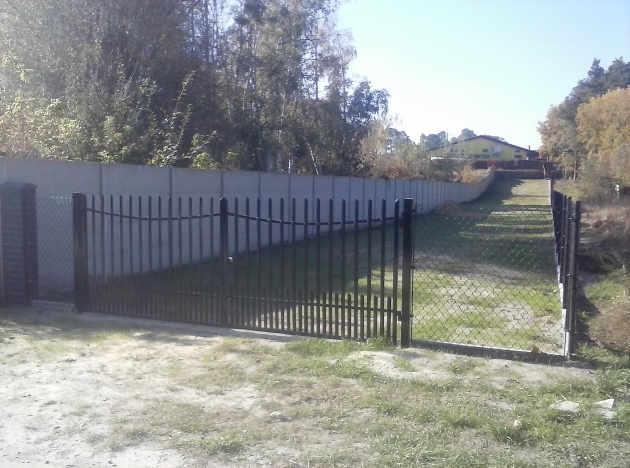 Nowoczesne ogrodzenie posesji w Sieciechowie: brama z metalowymi sztachetami, siatka ogrodzeniowa i wysoki betonowy mur oddzielający teren od drogi.