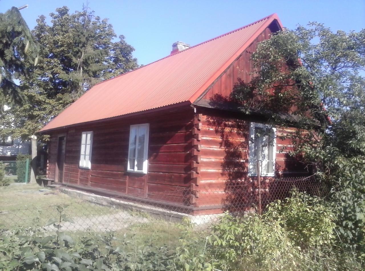 Czerwony, drewniany dom z blachodachówką, widoczny zza metalowej siatki ogrodzeniowej i bujnej roślinności.