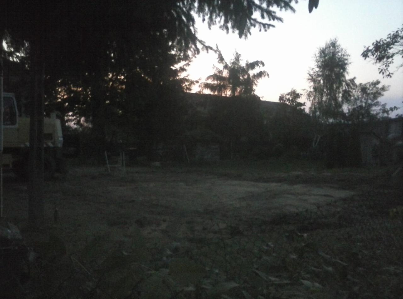 Zacieniony teren z przygotowaną ziemią, widoczne drzewa i sprzęt budowlany w tle, o zmierzchu.