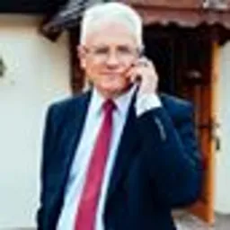 Mężczyzna w garniturze z czerwoną kravatą rozmawia przez telefon komórkowy na tle jasnego budynku.