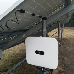 Widok od dołu na panele słoneczne zamontowane na metalowej konstrukcji, z widocznym białym falownikiem Huawei przymocowanym do słupka, okablowanie i trawa w tle.