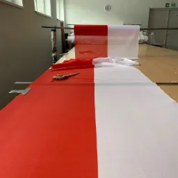 Czerwono-biały materiał rozłożony na stole w szwalni, nożyk krawiecki i metalowy docisk widoczne na czerwonej części materiału, w tle rolki materiałów i okno.