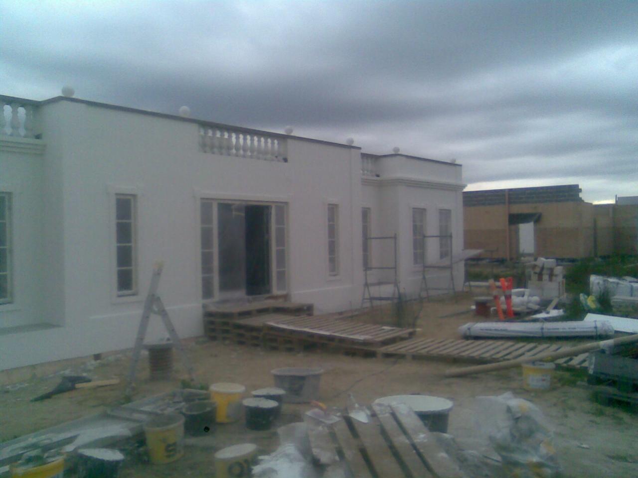 Biały budynek w trakcie wykończenia elewacji, widoczne rusztowania, palety i pojemniki z farbą, pochmurne niebo w tle.