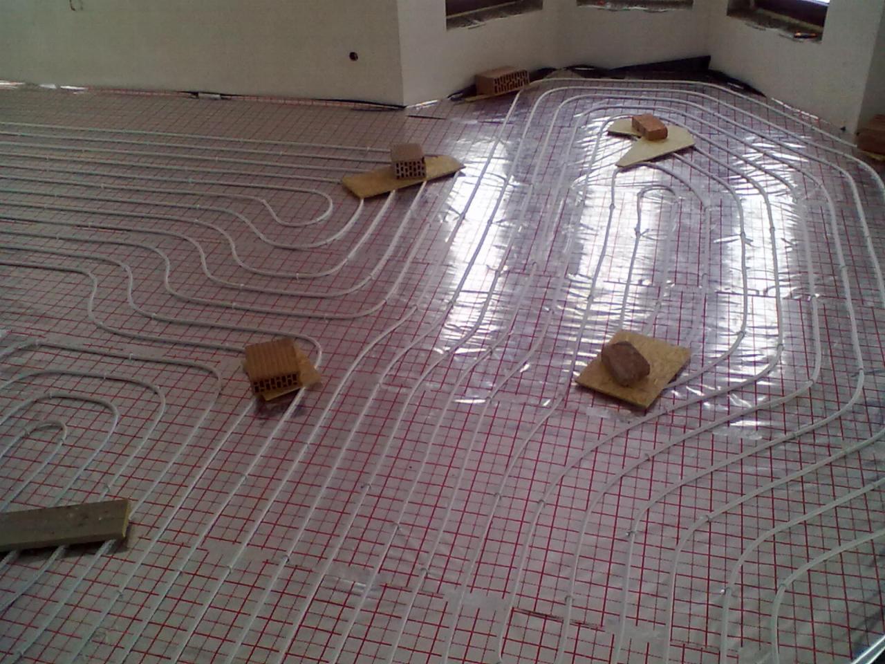 Białe rurki ogrzewania podłogowego ułożone na folii z czerwoną siatką, przytrzymywane cegłami i kawałkami drewna podczas montażu.