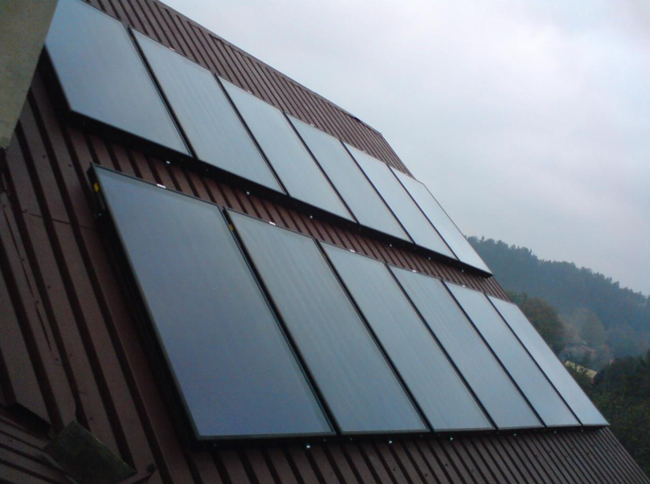 Rząd paneli solarnych zamontowanych na brązowym, skośnym dachu budynku, widok z dołu pod kątem, zalesione wzgórze w tle pod pochmurnym niebem.