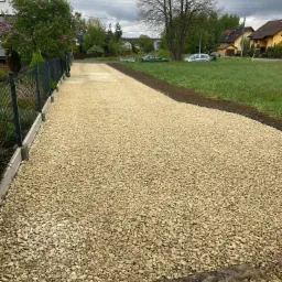Gotowa droga dojazdowa z jasnego kruszywa, obrzeża betonowe i trawnik. Widok z perspektywy pieszego. W tle zabudowa jednorodzinna i drzewa.