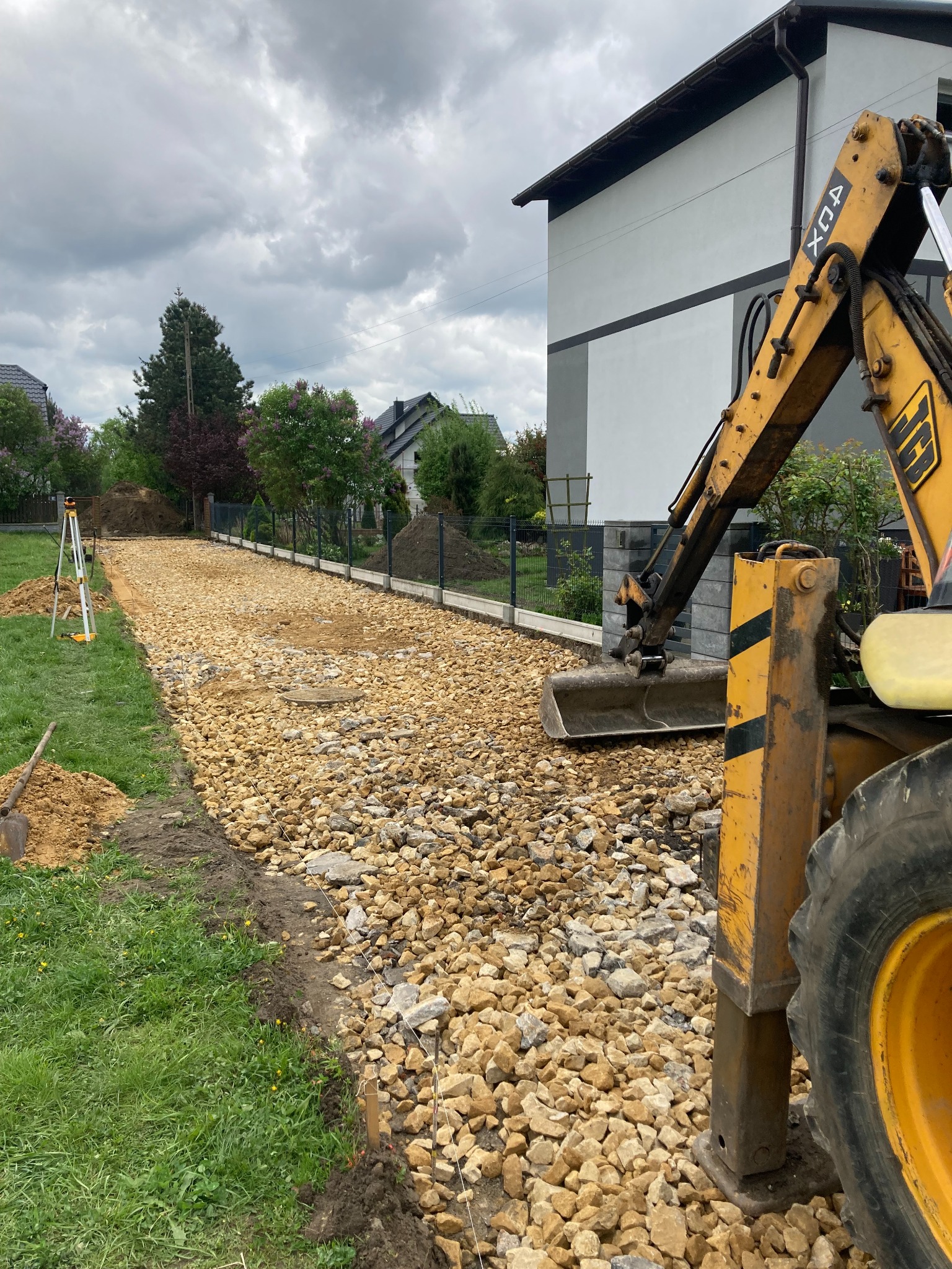 Budowa drogi dojazdowej z użyciem koparki na posesji z ogrodzeniem. Widoczne kamienie, ziemia i dom w tle. Prace w toku, teren wyrównany.