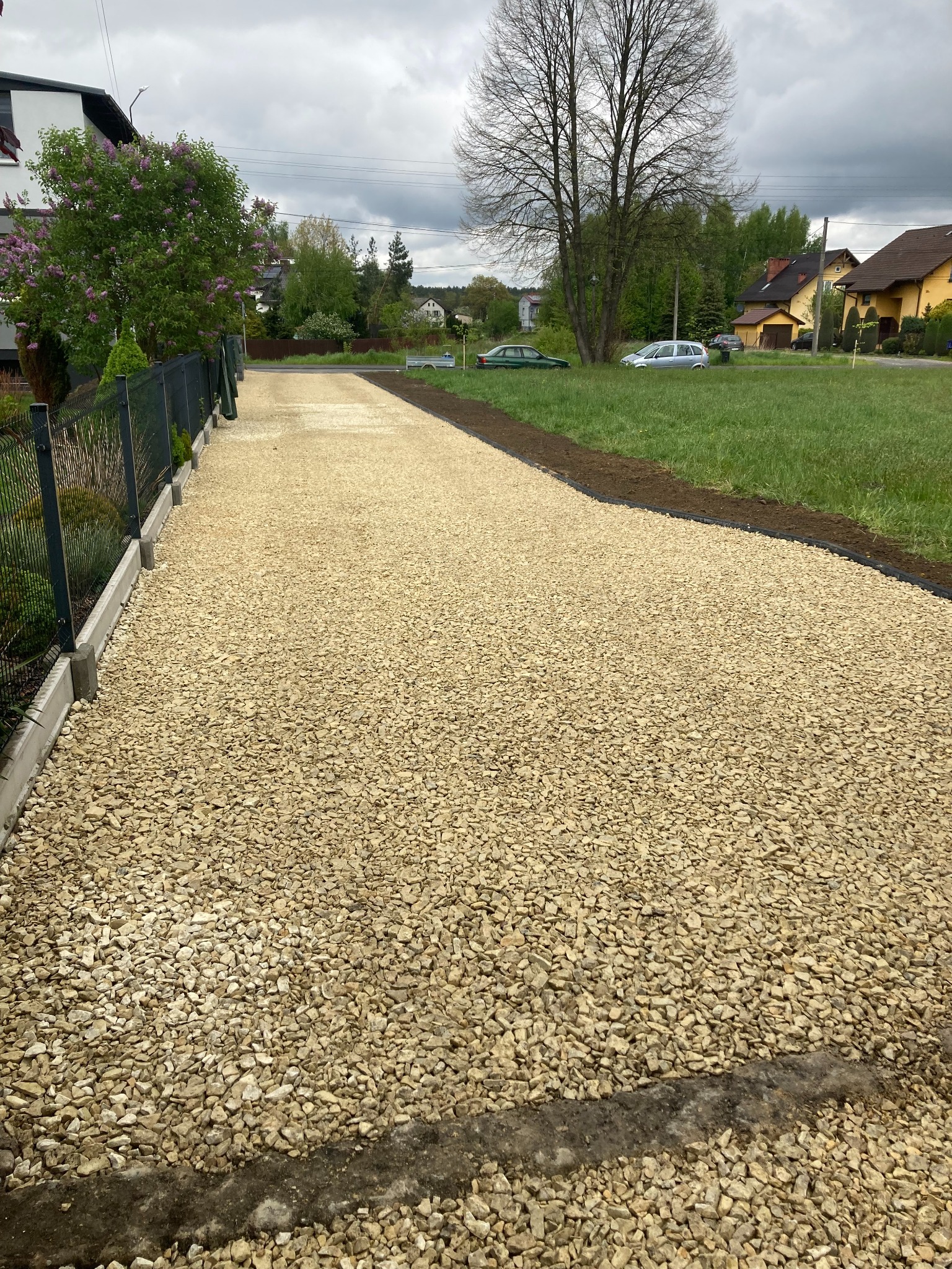 Gotowa droga dojazdowa z jasnego kruszywa, obrzeża betonowe i trawnik. Widok z perspektywy pieszego. W tle zabudowa jednorodzinna i drzewa.