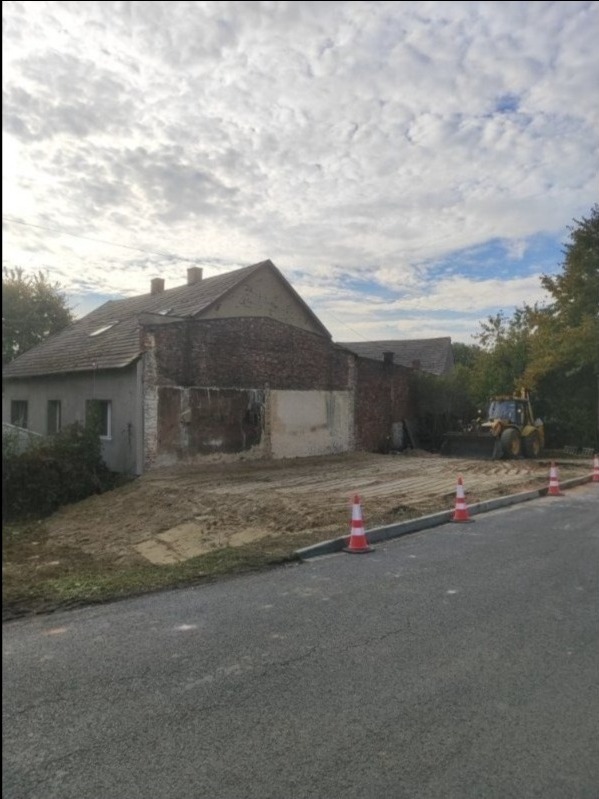 Częściowo rozebrany budynek z odsłoniętą cegłą, przygotowany teren, pomarańczowe pachołki i koparka w tle. Widok z poziomu ulicy.
