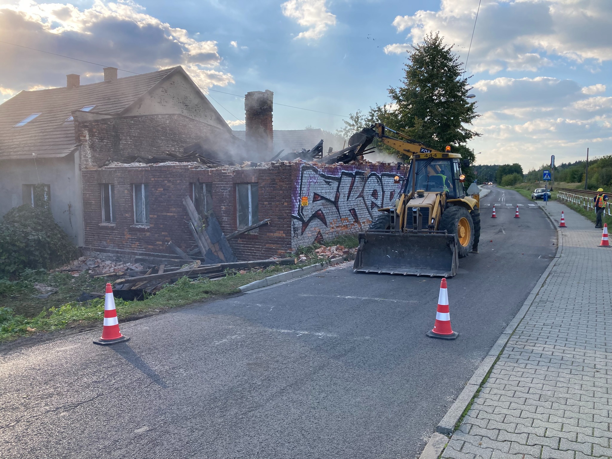 Żółta koparko-ładowarka na drodze, w tle częściowo zburzony budynek z cegły z graffiti, unoszący się dym, stożki ostrzegawcze. Prace rozbiórkowe w toku.