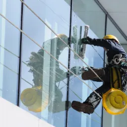 Pracownik w kasku i uprzęży, zawieszony na linach, myje szklaną fasadę budynku za pomocą myjki z długim uchwytem; widoczny żółty kubeł i odbicie pracownika w szybie.