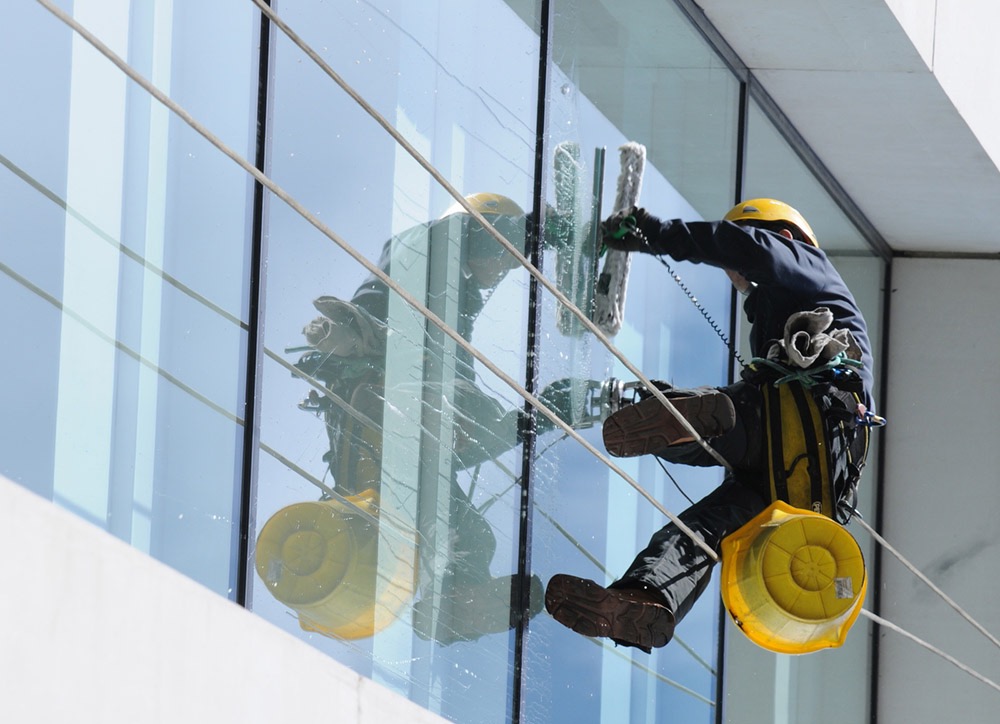 Pracownik w kasku i uprzęży, zawieszony na linach, myje szklaną fasadę budynku za pomocą myjki z długim uchwytem; widoczny żółty kubeł i odbicie pracownika w szybie.