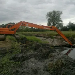 Pomarańczowa koparka gąsienicowa Doosan z długim ramieniem pracuje w korycie rzeki, pogłębiając dno.